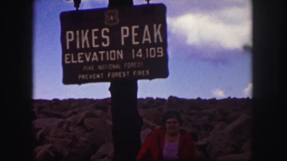 1958 Pikes Peak home-movie still showing the summit elevation sign, rugged rocks, and a woman posed beneath it.