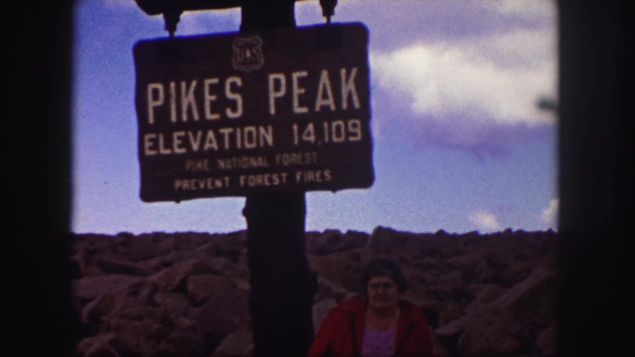1958 Pikes Peak summit view with the elevation sign centered, clouds overhead, and a visitor partly visible at the lower edge.