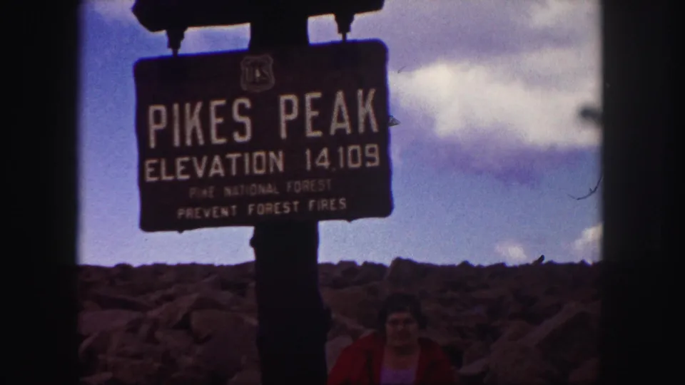 Color 8mm frame of the Pikes Peak elevation sign against rocks and sky, with a red-jacketed visitor partly visible below.