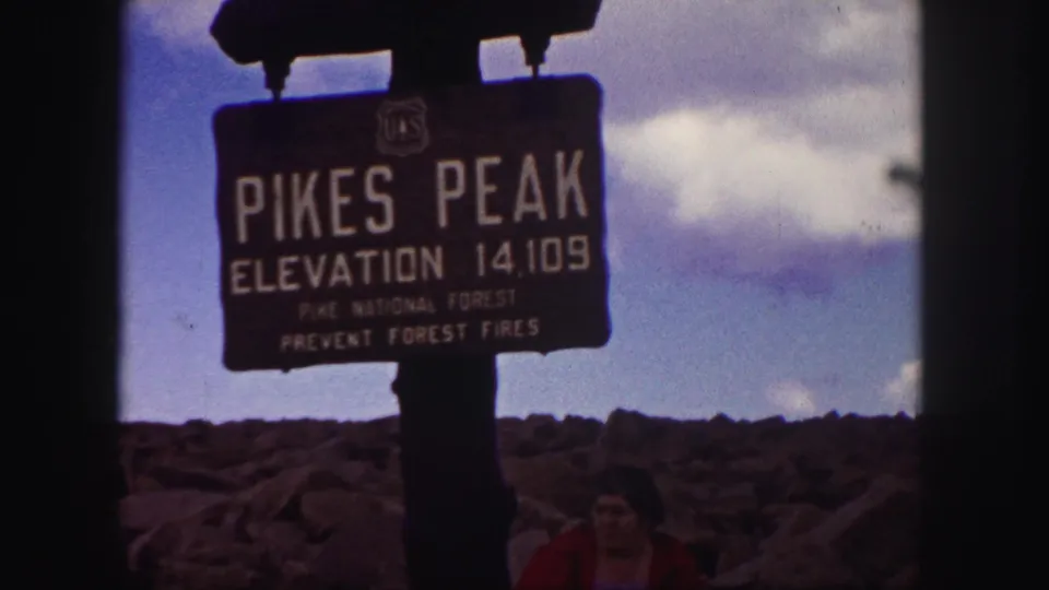 1958 Colorado Springs area home-movie frame showing the Pikes Peak sign, rocky summit foreground, clouds, and a visitor looking downward.