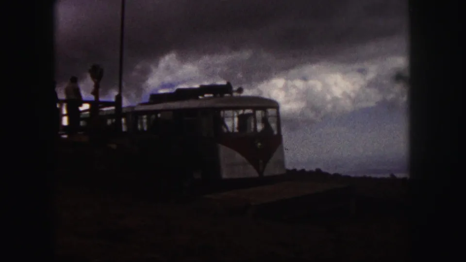 1958 Pikes Peak footage showing a red-and-white tourist vehicle near a platform with visitors under a dramatic cloudy sky.