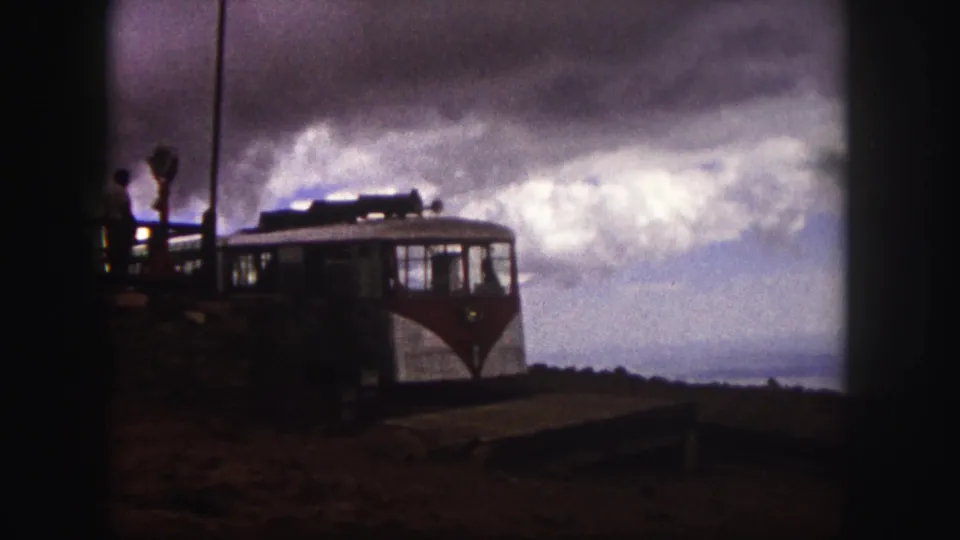 The tourist vehicle is seen from a handheld 8mm viewpoint, with visitors at the platform and clouds over the mountain landscape.