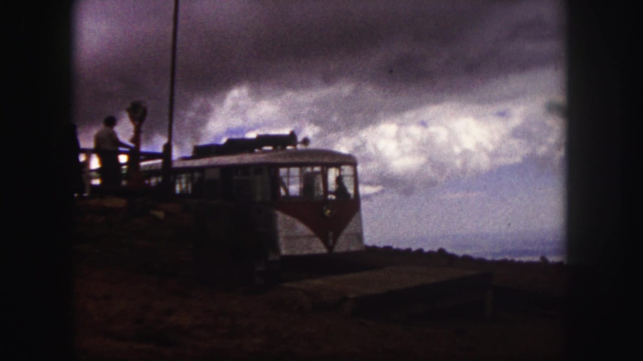 1958 Colorado Springs home-movie frame showing a red-and-white mountain train near a platform with passengers silhouetted by cloudy sky.