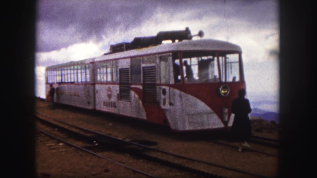 1958 Colorado Springs home-movie frame showing the side and front of a red-and-white mountain train stopped on rails with a passenger nearby.