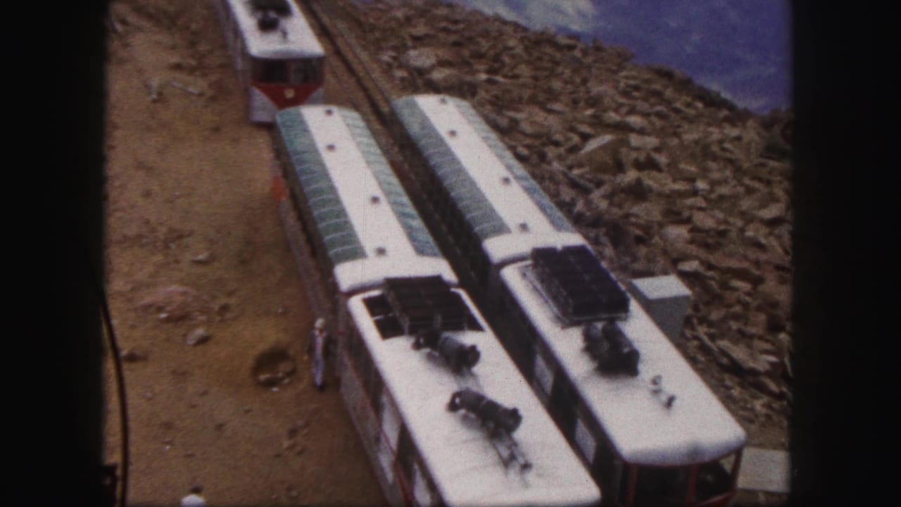 1958 Colorado Springs home-movie frame showing red-and-white rail cars on a rocky mountain railway with a small trackside figure and distant slopes.