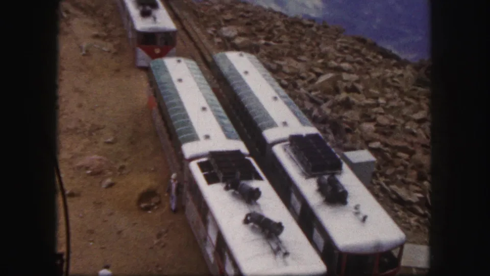 1958 Colorado Springs home-movie frame showing red-and-white rail cars on a rocky mountain railway with a small trackside figure and distant slopes.
