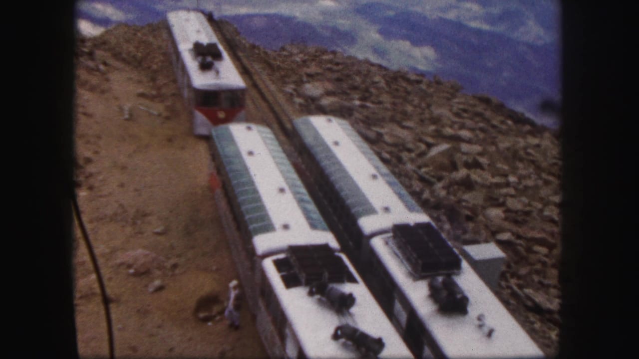 Mountain rail cars seen from above beside rocky ground and a cloudy mountain view.