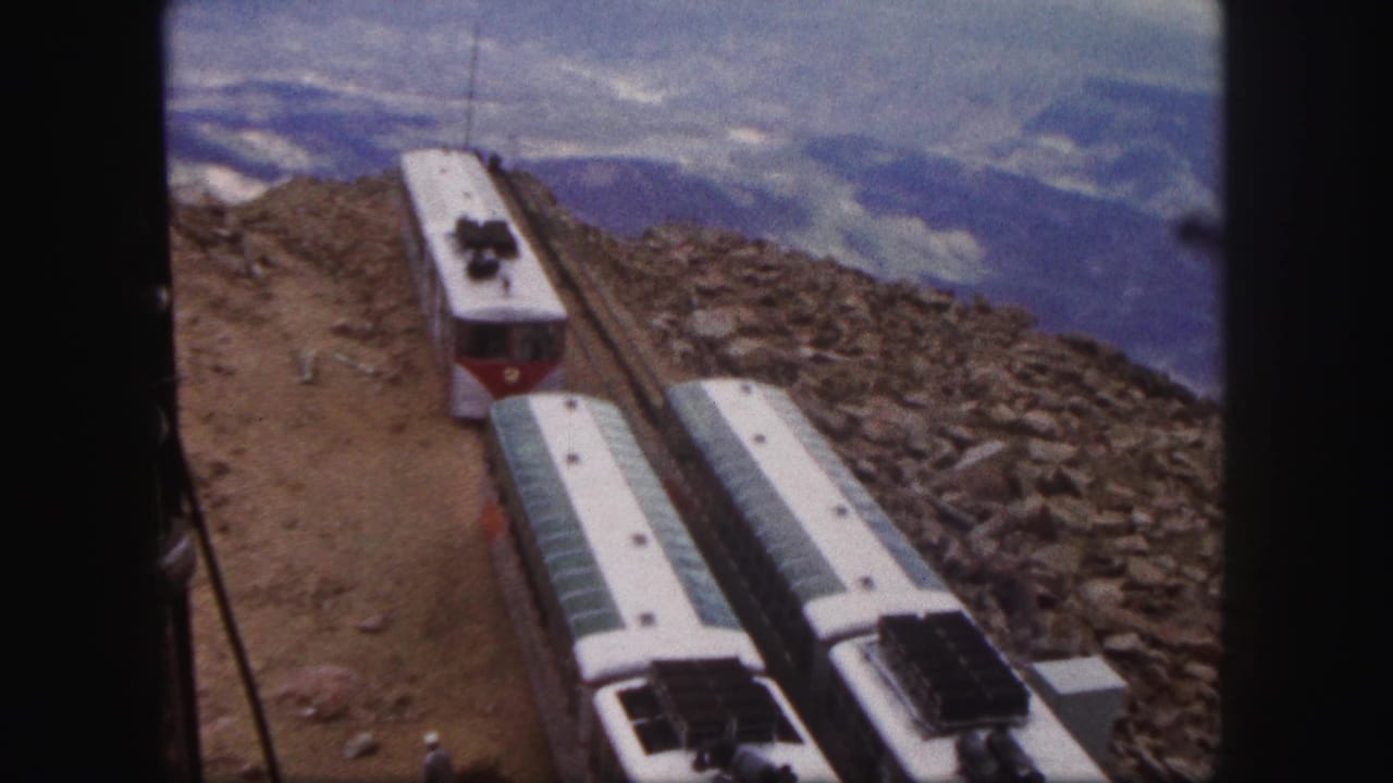1958 Colorado Springs 8mm frame showing red-and-white mountain railway cars on a steep rocky route with hazy mountains in the background.