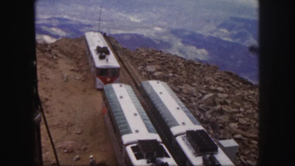 1958 Colorado Springs 8mm frame showing red-and-white mountain railway cars on a steep rocky route with hazy mountains in the background.