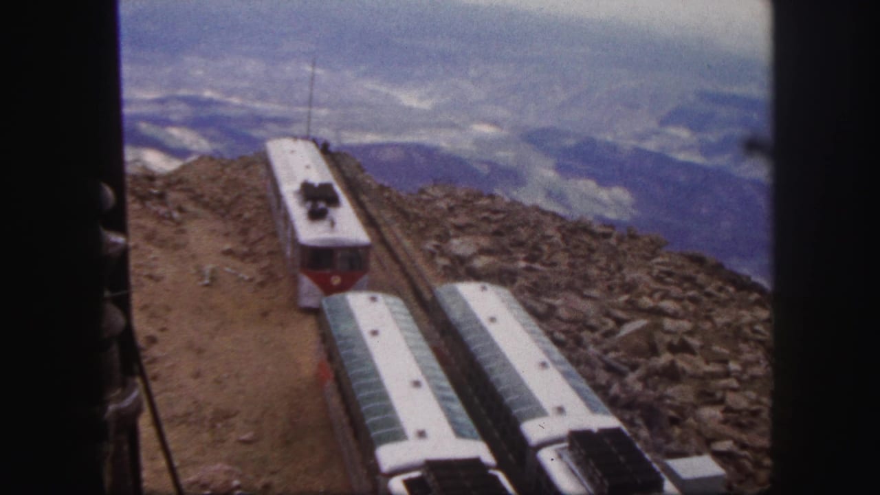1958 Colorado Springs frame of mountain railway cars on exposed rocky track, with distant blue mountain ridges and cloudy sky beyond.