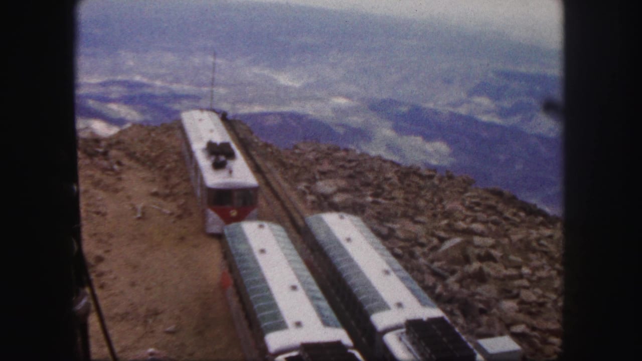 1958 Colorado Springs home-movie frame showing red and white railway cars on an elevated rocky mountain track under cloudy sky.