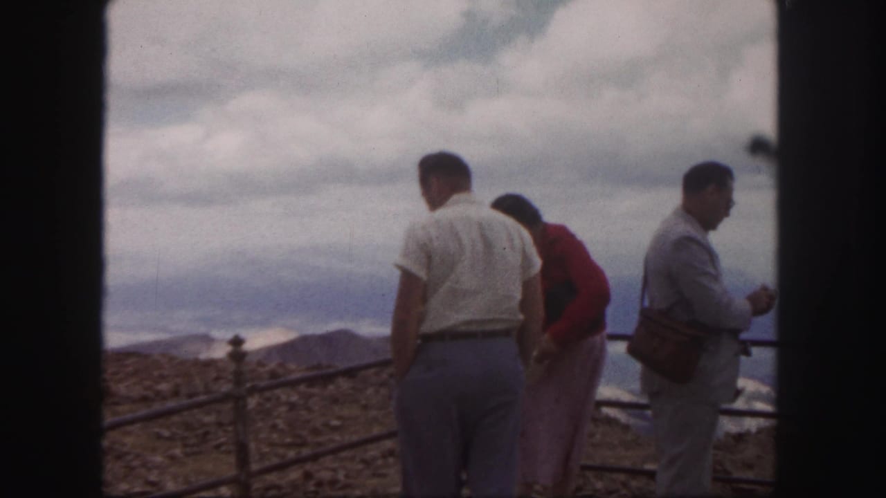 Three adult tourists stand by a metal guardrail at a mountain overlook.