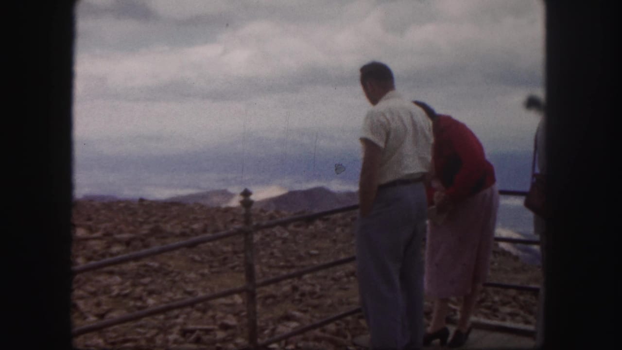 1958 Colorado Springs home-movie frame of visitors beside a metal overlook rail with rocky terrain and distant ridges behind them.
