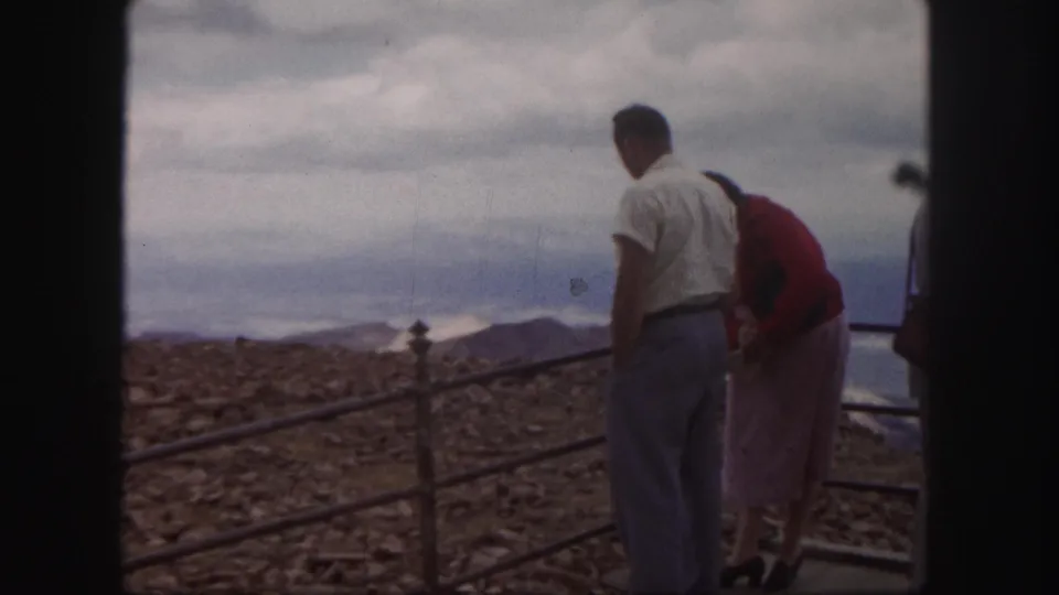 1958 Colorado Springs home-movie frame of visitors beside a metal overlook rail with rocky terrain and distant ridges behind them.
