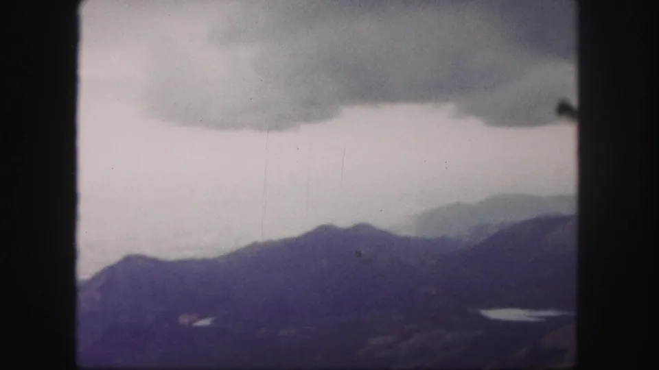 1958 8mm view from Pikes Peak showing layered Colorado mountain ridges, cloud cover, and a small bright lake in the distance.