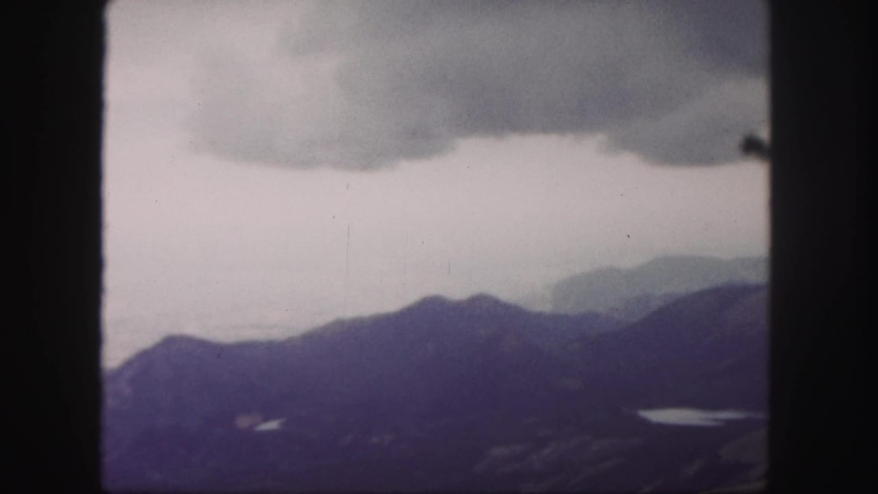1958 8mm landscape view from Pikes Peak with stormy clouds, shadowed mountain ridges, and a small reflective lake below.