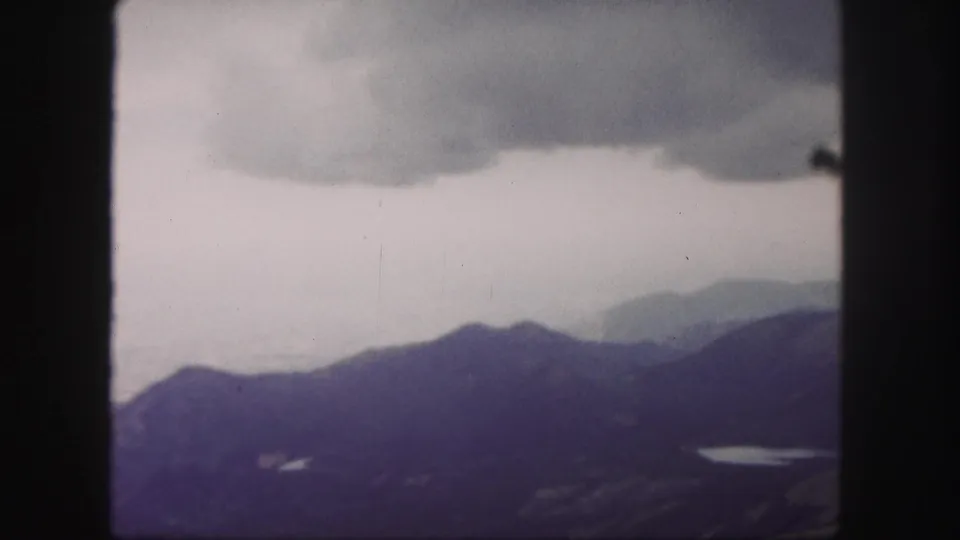 1958 8mm landscape view from Pikes Peak with stormy clouds, shadowed mountain ridges, and a small reflective lake below.