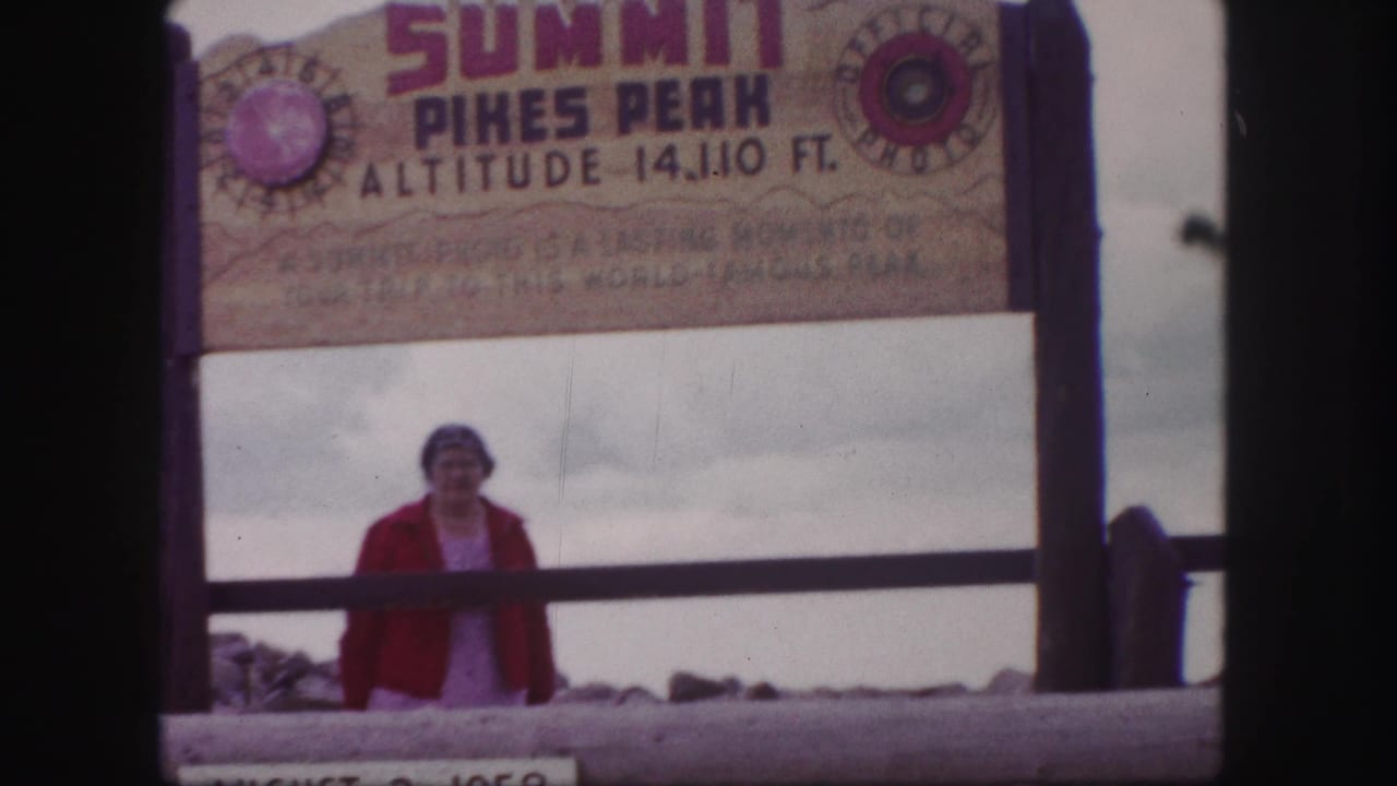 Woman in a red jacket poses beneath the Summit Pikes Peak sign marked 14,110 feet, with cloudy sky and summit railing behind her.