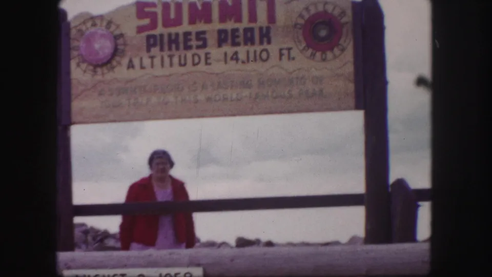 Woman in a red jacket poses beneath the Summit Pikes Peak sign marked 14,110 feet, with cloudy sky and summit railing behind her.