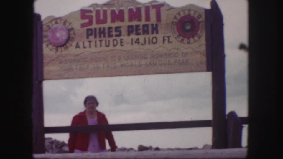 1958 Colorado Springs home-movie frame showing a woman below the Pikes Peak summit altitude sign.