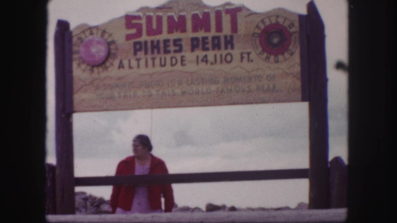 Home-movie frame of a woman glancing to the side beneath the Pikes Peak summit sign in Colorado.