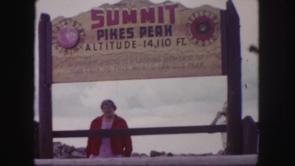 1958 Pikes Peak summit frame showing the woman under the sign and a partial figure entering at right.