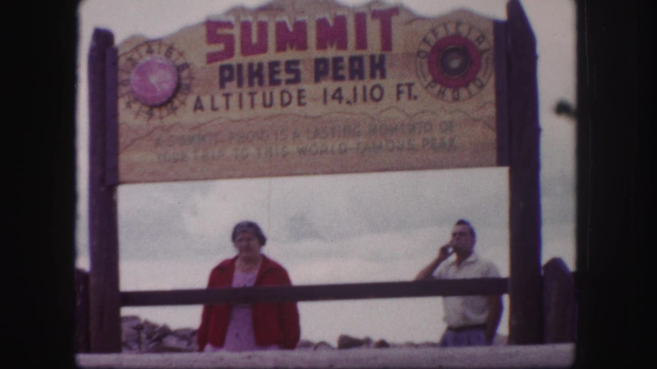 Two adults stand beneath the Pikes Peak summit sign with a wooden railing in front.