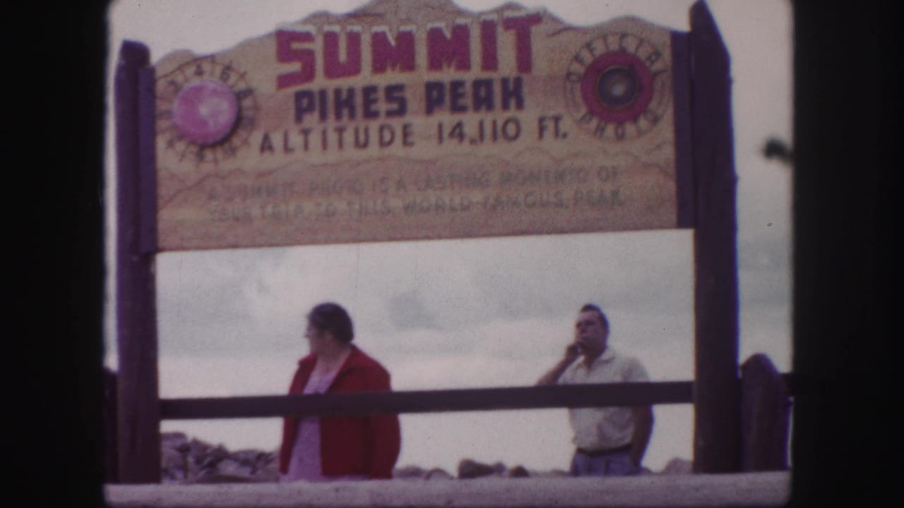 Two adults beneath the Pikes Peak summit sign, with one person turned sideways.