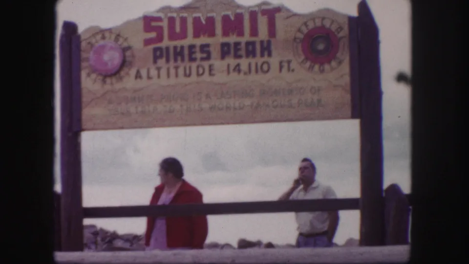 Colorado Pikes Peak summit view with two adult visitors under the wooden altitude sign, filmed on 8mm in 1958.
