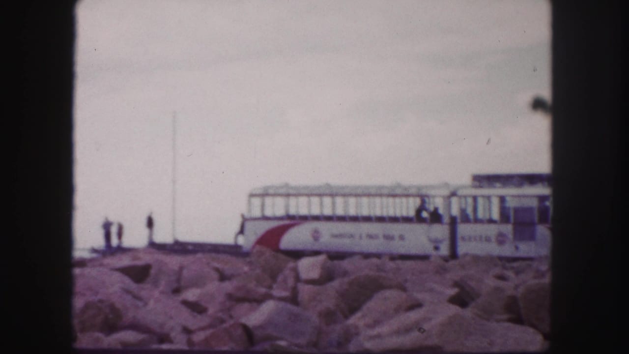 1958 8mm view of a Pikes Peak railway car on the rocky summit area, with small figures nearby and cloudy sky.