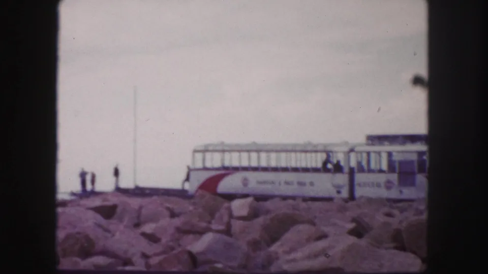 1958 8mm view of a Pikes Peak railway car on the rocky summit area, with small figures nearby and cloudy sky.