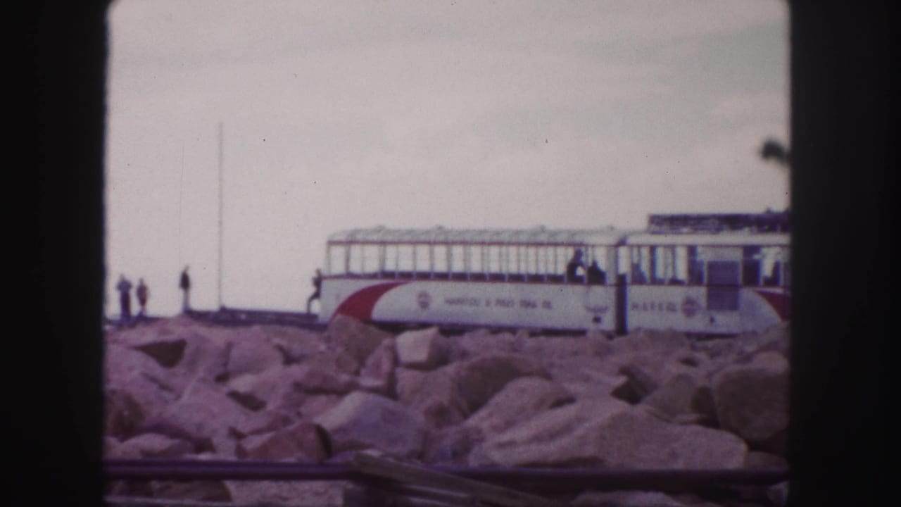 The camera holds on the Pikes Peak Cog Railway car across foreground rocks, with visitors silhouetted near the track.