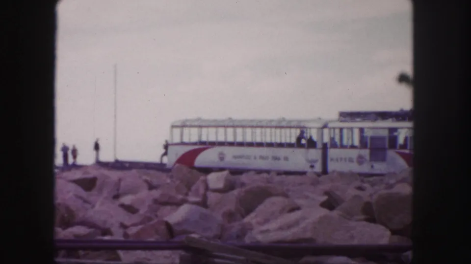 The camera holds on the Pikes Peak Cog Railway car across foreground rocks, with visitors silhouetted near the track.