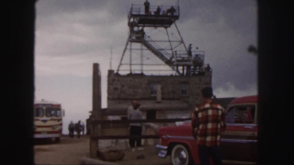 A summit-area stone building and metal observation tower are shown with visitors, a vintage bus, and a red car in 1958 footage.