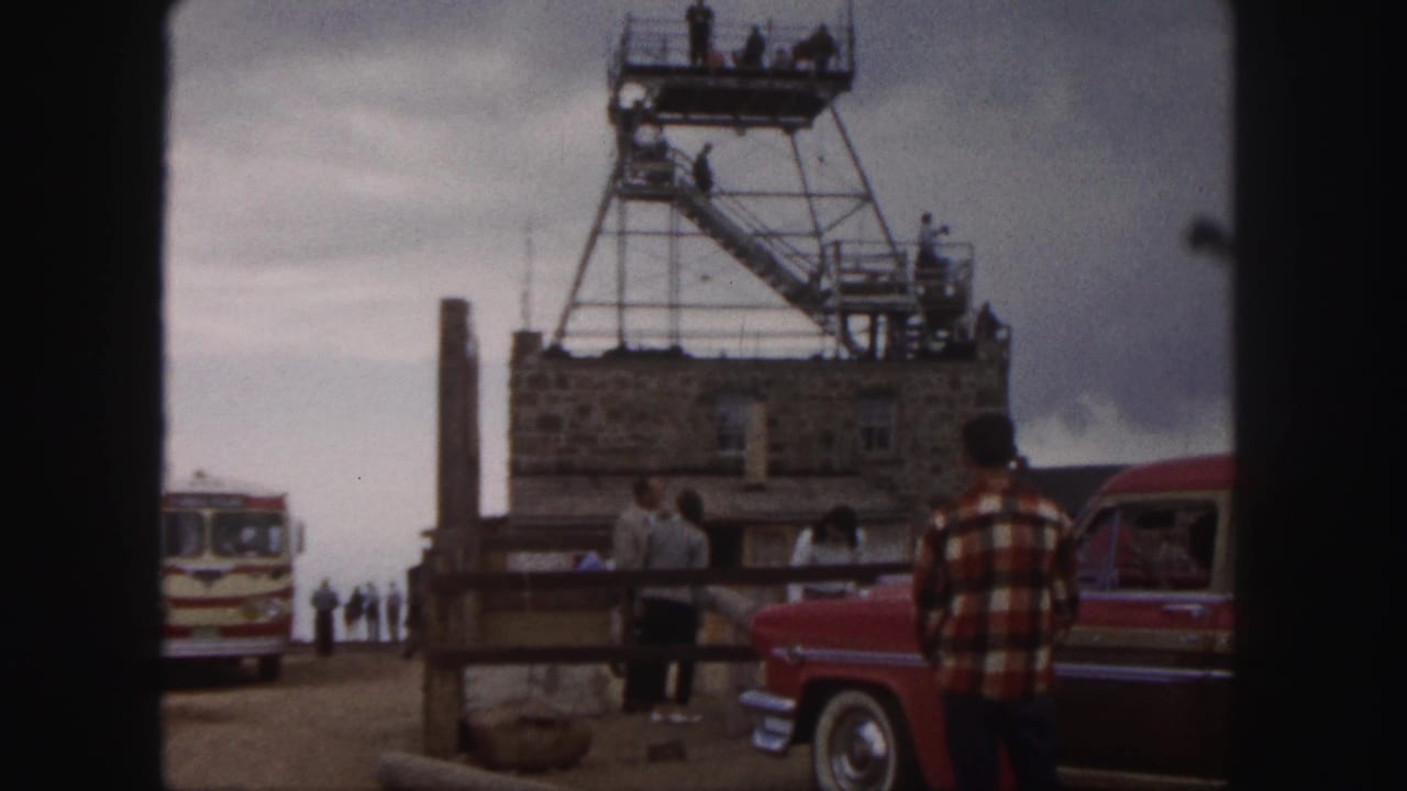 1958 Colorado Springs home-movie frame showing visitors by a stone overlook building, tower stairs, red car, and striped tour bus.