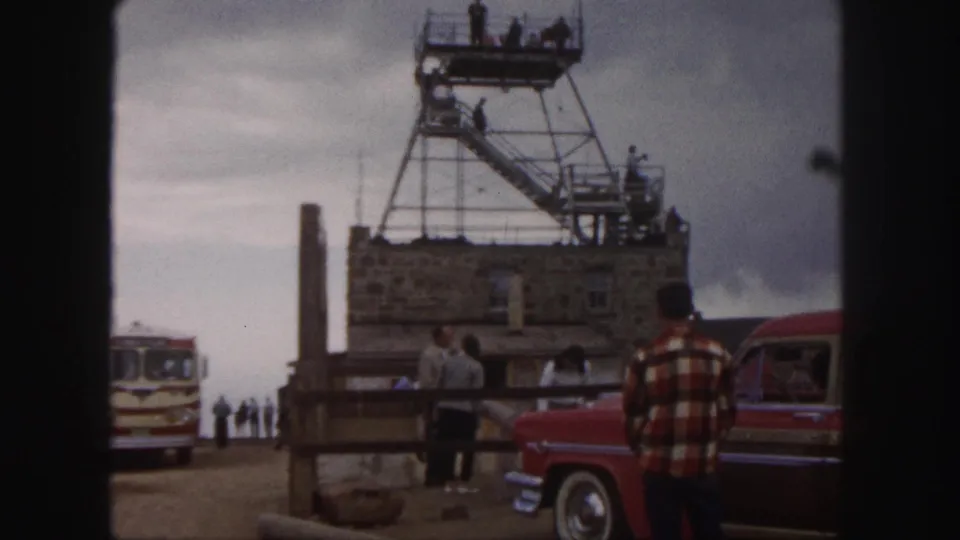 1958 Colorado Springs home-movie frame showing visitors by a stone overlook building, tower stairs, red car, and striped tour bus.