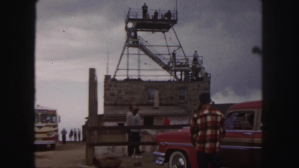1958 Colorado Springs overlook view with visitors near a stone structure, metal tower stairs, red car, and tour bus.