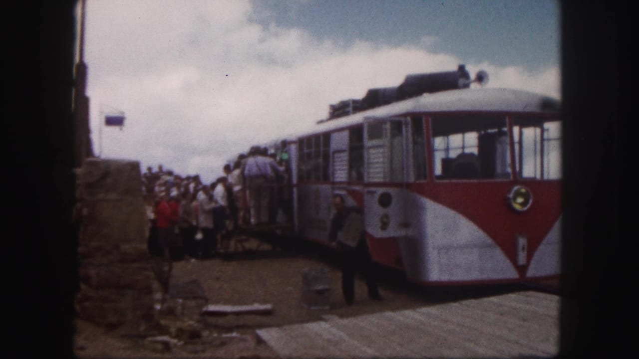 A red and white car waits as tourists queue along the platform beside stonework.