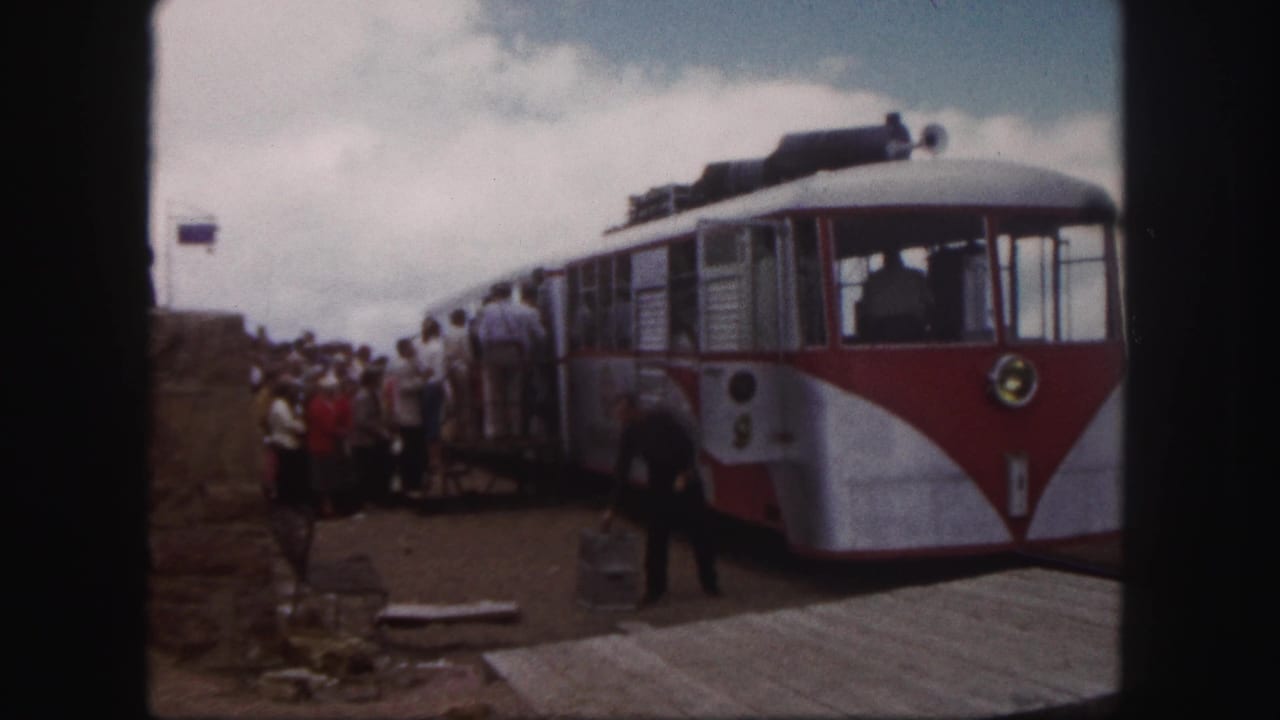 1958 Colorado Springs frame showing tourists boarding or waiting beside a red and white mountain transport car at a station platform.