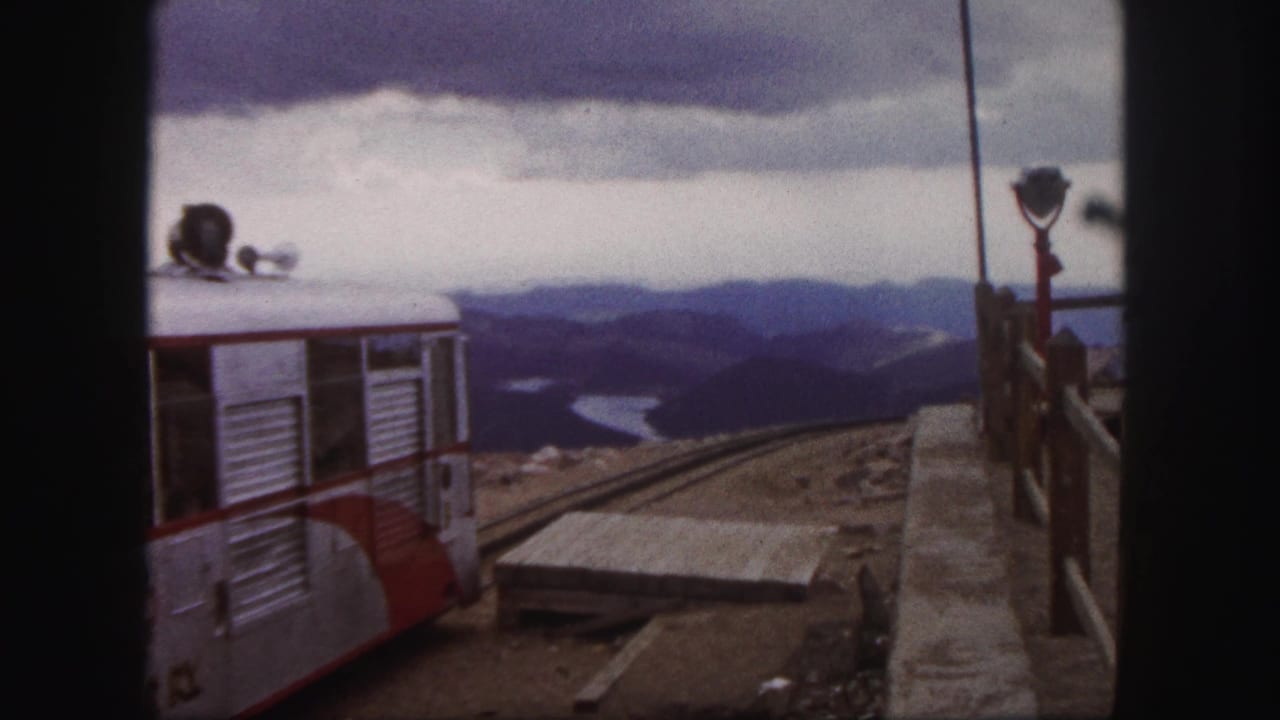 1958 Colorado Springs 8mm frame showing a red and silver tour train at a mountain station with distant ridges and water below.