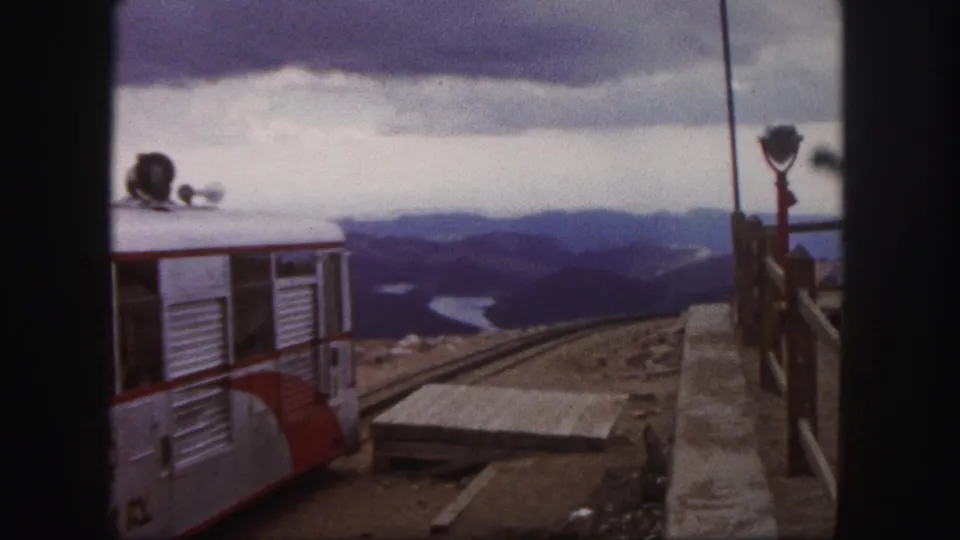 1958 Colorado Springs 8mm frame showing a red and silver tour train at a mountain station with distant ridges and water below.