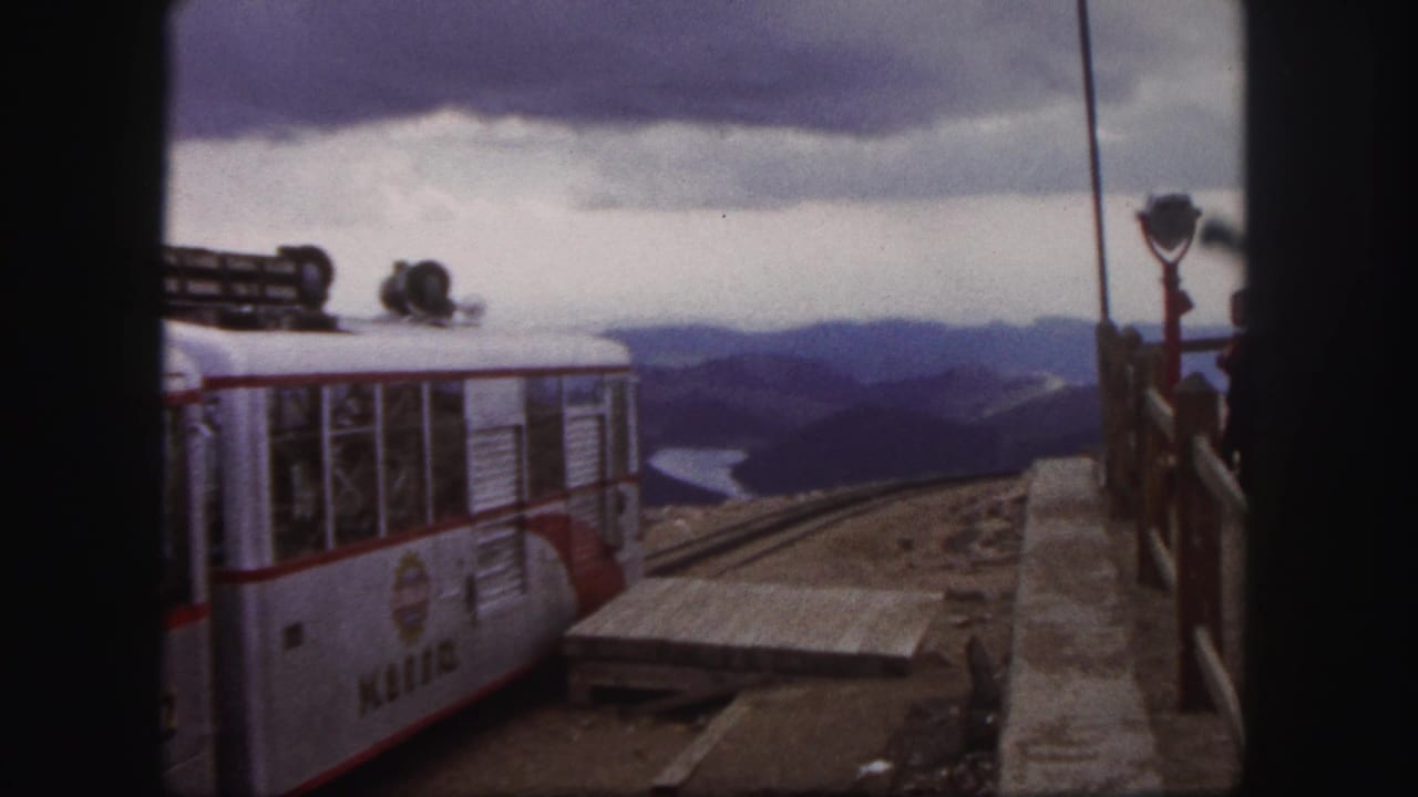 Tour train car passing a platform with mountain scenery in the background.
