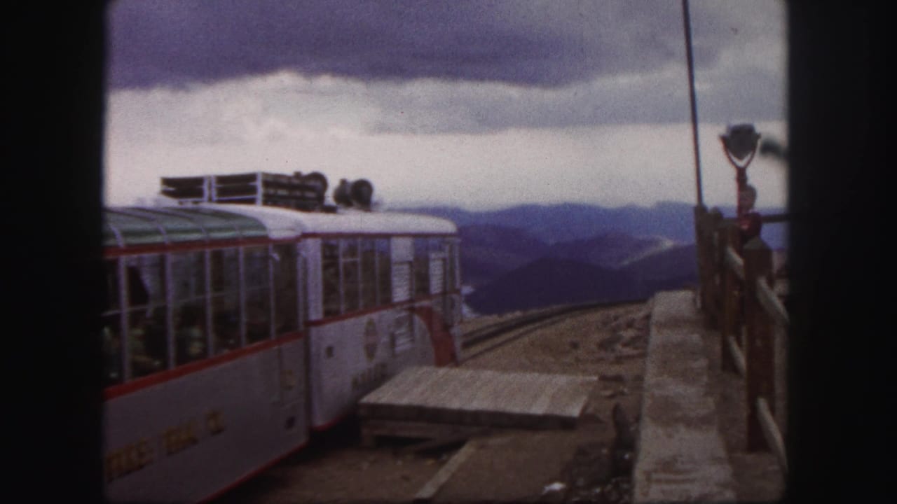1950s home-movie frame of a red-trimmed silver tour train departing a mountain station, with passengers faintly visible through windows.