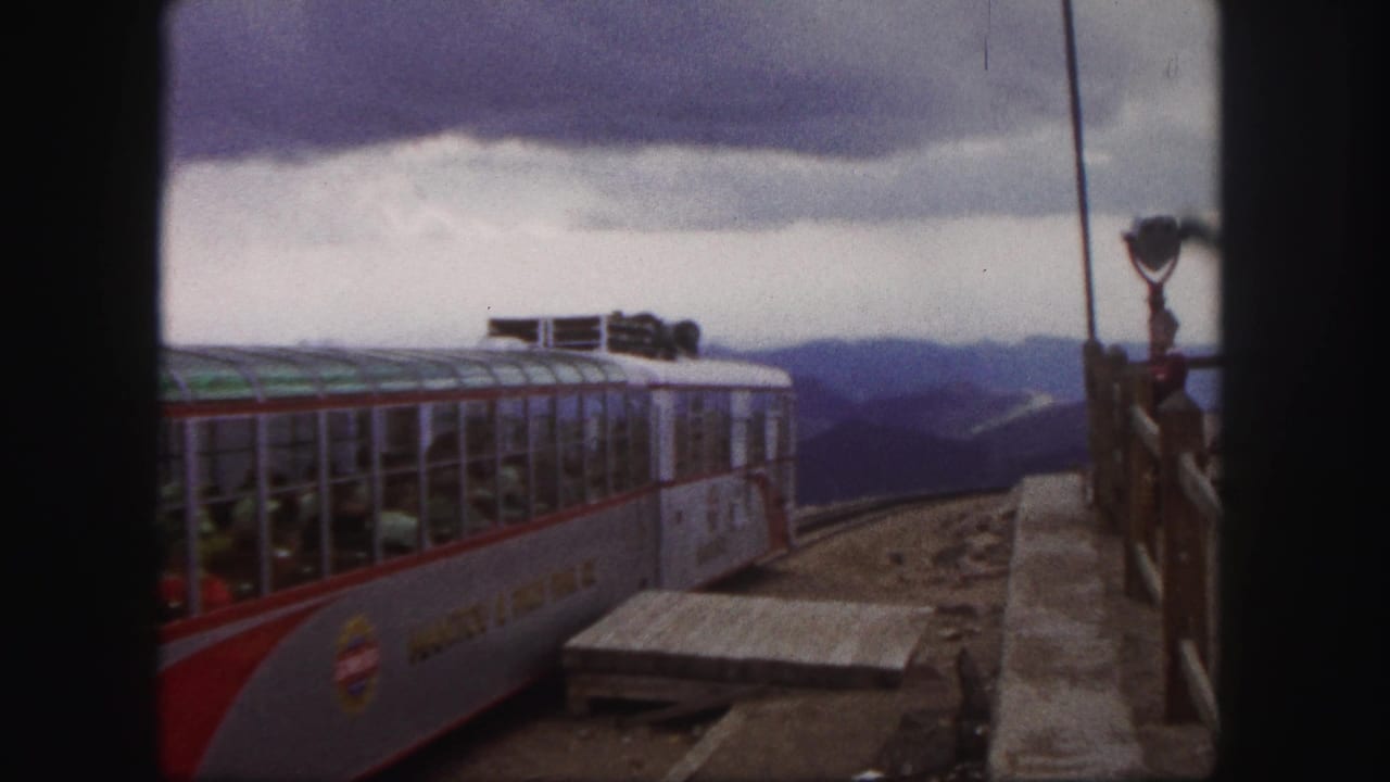 Colorado Springs tour train continues its departure in a 1958 8mm frame, with passengers, railings, mountains, and cloudy sky visible.