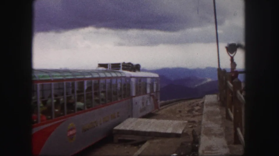Colorado Springs tour train continues its departure in a 1958 8mm frame, with passengers, railings, mountains, and cloudy sky visible.