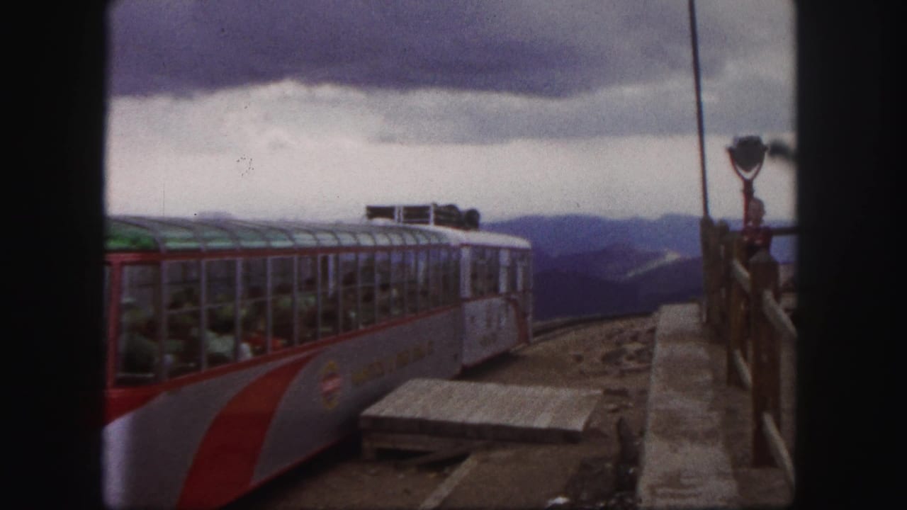 1958 Colorado Springs 8mm frame showing a red-and-white mountain railcar near a station platform under a cloudy mountain sky.