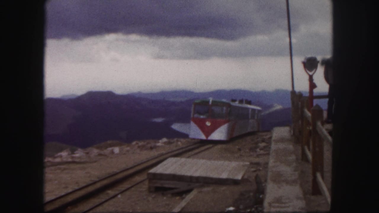 1958 Colorado Springs frame with the mountain passenger train farther along the track, overlook railings at right, and a lake in the distance.