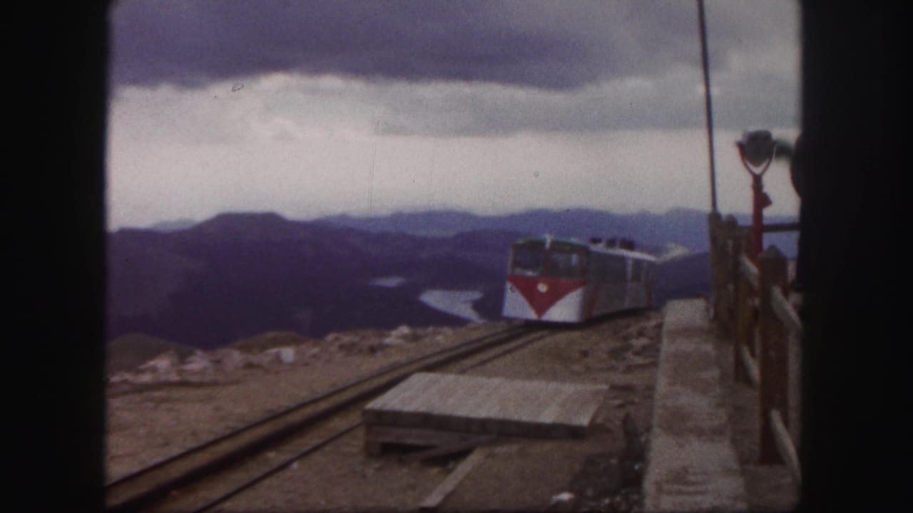1958 Colorado Springs home-movie still showing the red-and-white train on elevated mountain tracks near a wooden platform and distant lake.