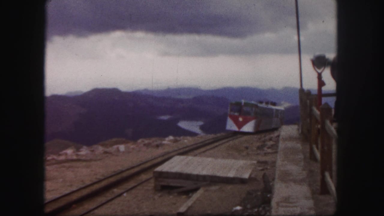 1958 Colorado Springs frame of a red-and-white passenger train moving out of view along mountain tracks, with lake, ridges, and overlook railings visible.