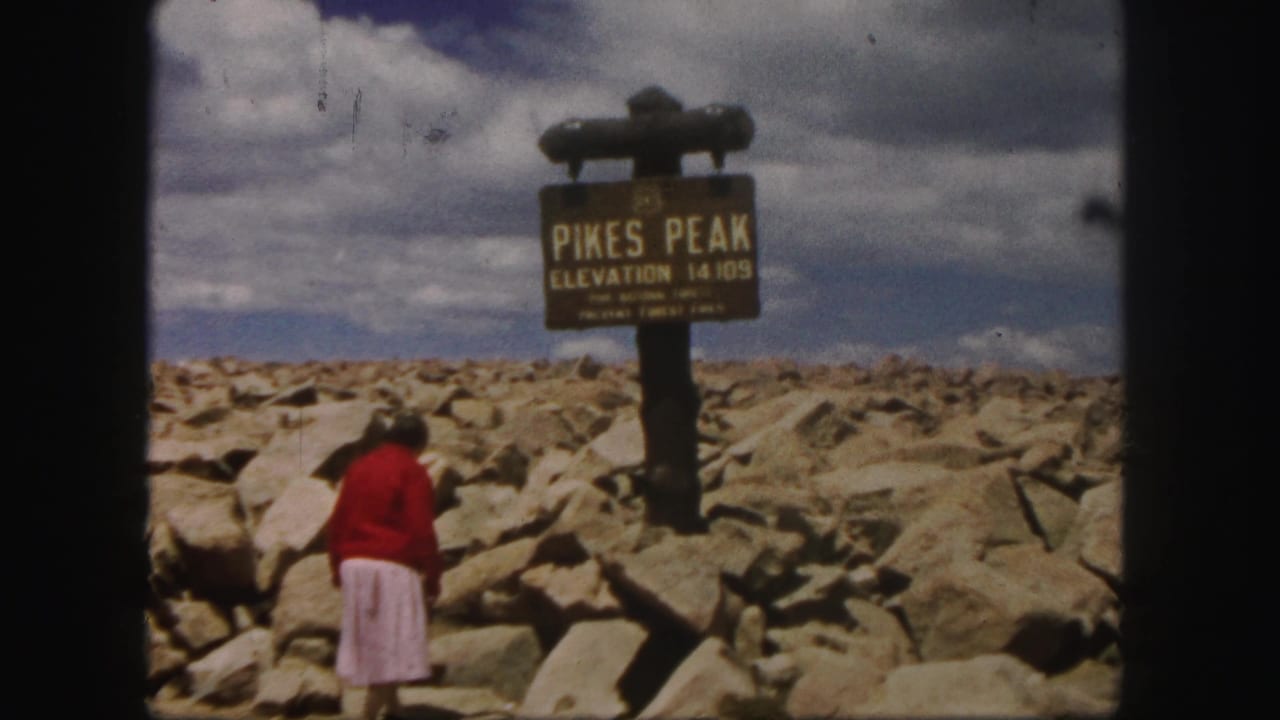 The woman moves closer to the Pikes Peak sign, with boulder-strewn summit terrain and a cloudy sky visible.